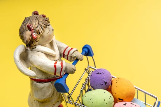 A sleepy Angel carries colorful Easter eggs in a shopping basket on a yellow background. space for copying. close-up. Easter greeting card.