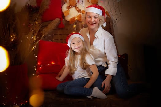 Cute mother and daughter in the red hats of Santa Claus assistants in a room decorated for Christmas. The tradition of decorating house and dressing up for holidays. Happy childhood and motherhood