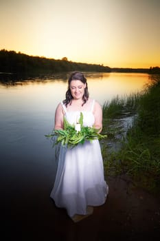 Slavic plump plump chubby girl in long white dress on the feast of Ivan Kupala with flowers and water in a river or lake on summer evening