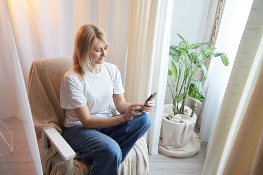 Young blonde woman using cell phone at home. Girl is resting, chating and looking in camera on smartphone near window at home. Nice atmosphere. The concept of cozy home and communication by Internet