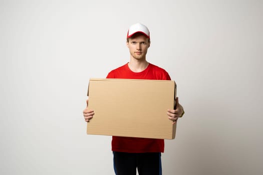 Portrait of a young bearded delivery man in red uniform holding blank cardboard, percel isolated on white background