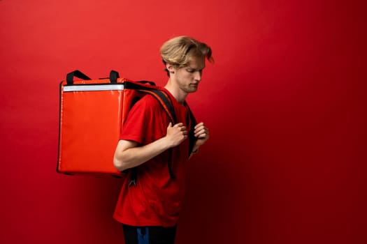 Side view of young courier with curly hair in red uniform with red thermo bag on red background. Food delivery service