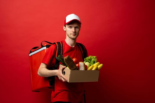 Proffesional young delivery man in red uniform carrying package box of grocery food and drink from store, market on red studio background. Delivery Concept