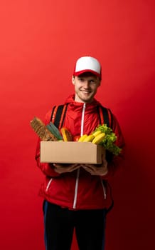 Fast young delivery man in red uniform carrying package box of grocery food and drink from store, market on red studio background. Delivery Concept