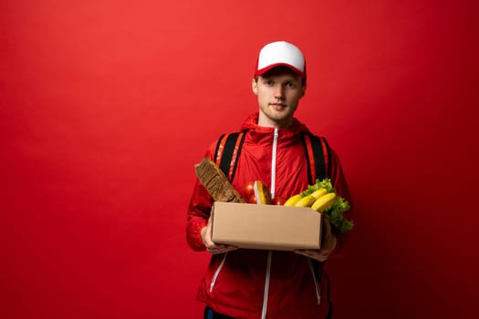 Young delivery man carrying paper box bag of grocery food and drink from store isolated on red studio background. Copy Space. Delivery Concept