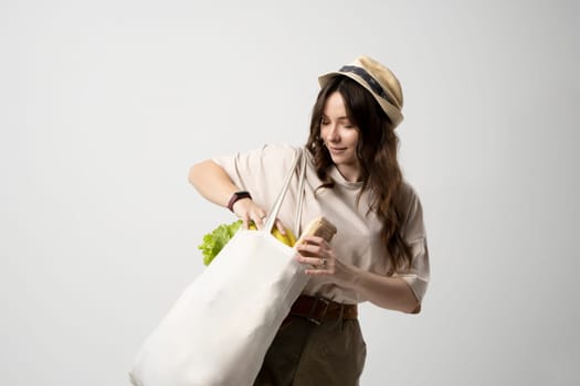 Smiling young woman in beige t-shirt and a hst with a mesh eco bag full of vegetables and on a white studio background. Sustainable lifestyle. Eco friendly concept. Zero waste