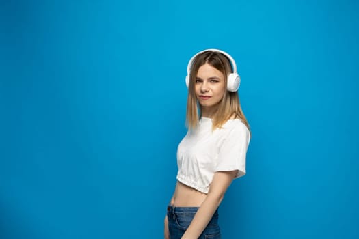 Cheerful young pretty girl smiling while listening music in headphones and dancing on blue background