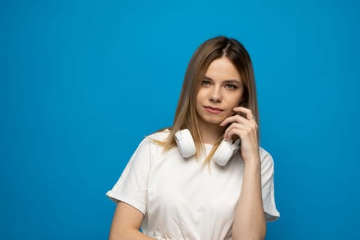 Young blonde beautiful girl in white outfit with a big headphones on a shoulder on blue background