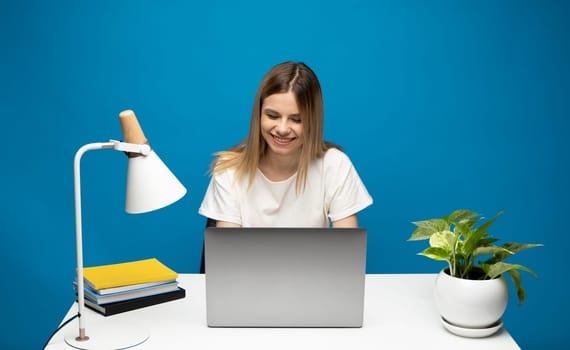 Portrait of a pretty young woman studying while sitting at the table with grey laptop computer, notebook. Smiling business woman working with a laptop isolated on a blue background