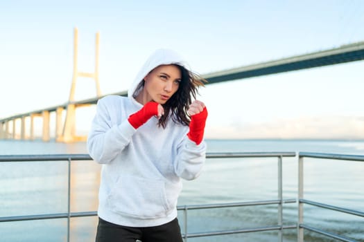 Woman shadow boxing with her hands wrapped in red boxing tapes with ocean bridge and blue sky background. Female athlete fighter training outside dressed hoodie. Copy space