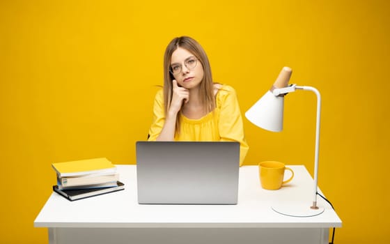 Portrait of Smiling pretty young woman studying while sitting at the table with grey laptop computer, notebook. Business woman working with a laptop isolated on a yellow background