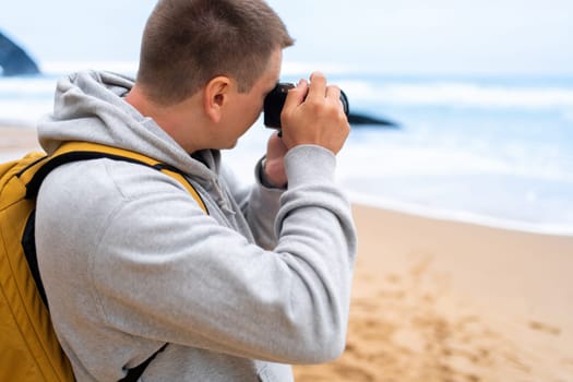 Traveler photographer takes photo beautiful seascape landscape on professional camera. Man tourist with backpack shoots a photo on beach. Side view middle shoot.