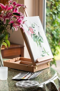 The easel with still life with purple lilies watercolor on the table prepared for painting at home
