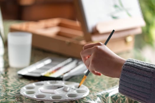 Young left-handed girl painting still life with flowers mixing watercolor paint in palette at home