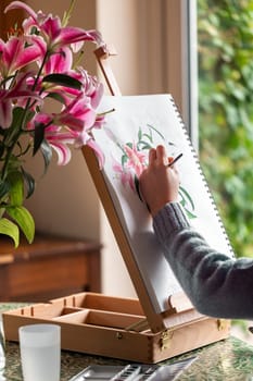 Young left-handed girl painting still life with flowers, purple lilies, with watercolor paints on the easel at home