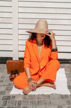 Stylish woman in an orange suit with a hat sits on a rug on a white striped background. On the hands are jewelry rings and bracelets, sandals and a bag stand side by side