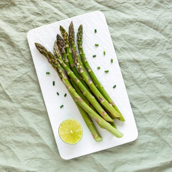 asparagus stems on the white rectangular plate served with green lemon and cut chives, top view, green linen background, square photo