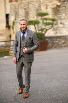 A man with a beard in a strict grey three-piece suit with a tie in the old town of Sirmione, a Stylish man in a grey suit in Italy.