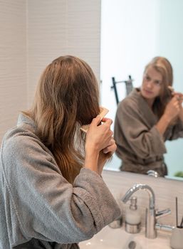 Womens health. Spa and wellness. Young woman in gray bathrobes brushing her hair and smiling while looking at the mirror
