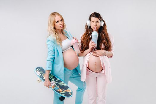 Two pregnant girls in turquoise and pink suits with glasses of juice, a skate and headphones stand on a gray background.