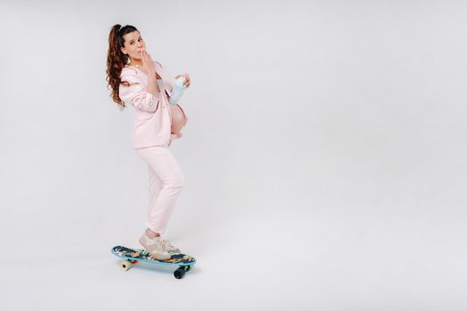 A pregnant girl in a pink suit with a skateboard in her hands stands on a gray background.