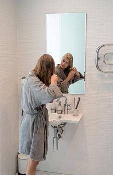 Womens health. Spa and wellness. Young woman in gray bathrobes brushing her hair and smiling while looking at the mirror