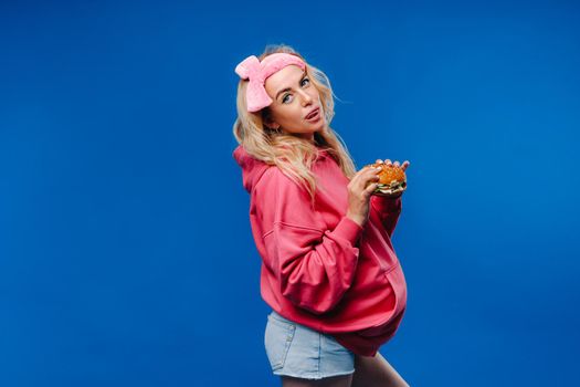a pregnant girl in pink clothes with a hamburger in her hands on a blue background.
