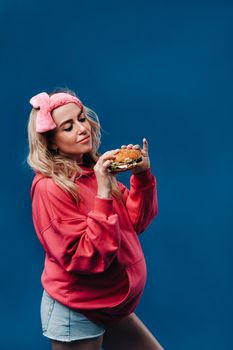 a pregnant girl in pink clothes with a hamburger in her hands on a blue background.