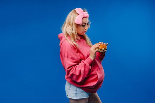 a pregnant girl in pink clothes with a hamburger in her hands on a blue background.