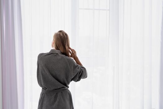 Womens health. Spa and wellness. Happy young woman applying hair mask standing next to the window. Rear view