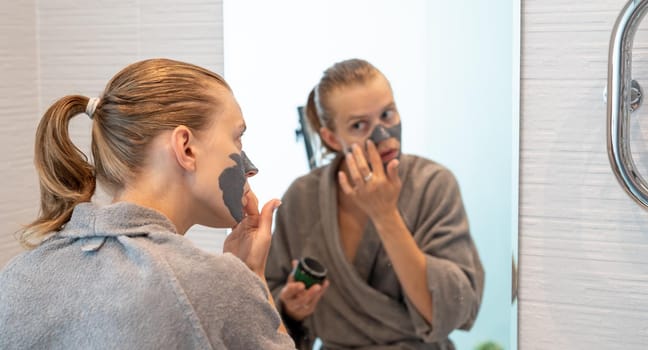 Womens health. Spa and wellness. Woman in gray bathrobes applying face mask in the bathroom looking at the mirror