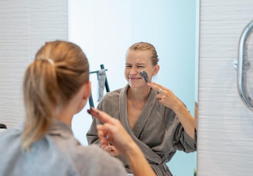 Womens health. Spa and wellness. Woman in gray bathrobes applying face mask in the bathroom looking at the mirror