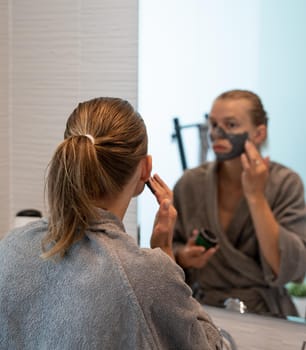 Womens health. Spa and wellness. Woman in gray bathrobes applying face mask in the bathroom looking at the mirror