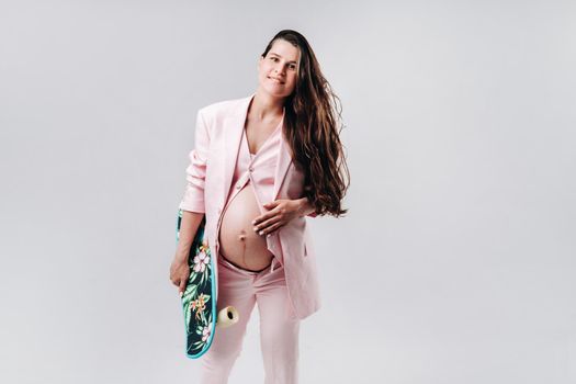 A pregnant girl in a pink suit with a skateboard in her hands stands on a gray background.