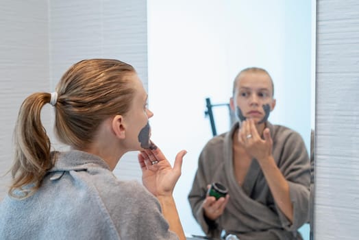 Womens health. Spa and wellness. Woman in gray bathrobes applying face mask in the bathroom looking at the mirror