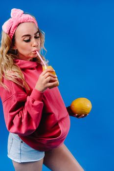 pregnant girl in pink clothes with a bottle of juice and a melon on a blue background.