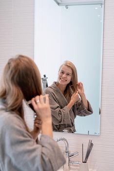 Womens health. Spa and wellness. Young woman in gray bathrobes brushing her hair and smiling while looking at the mirror