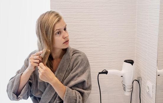 Womens health. Spa and wellness. Young woman in gray bathrobes brushing her hair and smiling while looking at the mirror