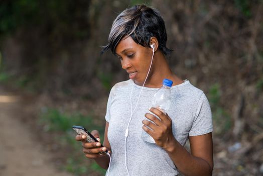 Slim woman athlete holding a bottle of water and using a modern cell phone while jogging outdoors. Sport concept