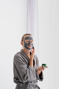 Womens health. Spa and wellness. Woman in gray bathrobes applying face mask in the bathroom looking at the mirror