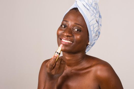 young woman on gray background with towel tied on her head putting on lipstick smiling.