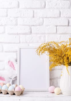 Spring holidays, Happpy Easter. Pastel colored Easter eggs and mimosa flowers with blank white frame for mockup design, front view on white brick wall background