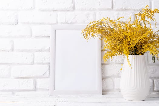 Spring holidays, Happpy Easter. Pastel colored Easter eggs and mimosa flowers with blank white frame for mockup design, front view on white brick wall background