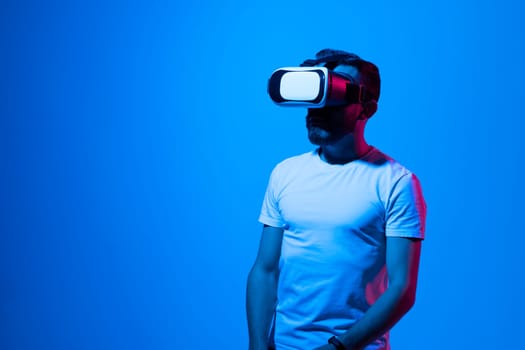 Bearded brunette man in white t-shirt play VR games in virtual reality helmets in neon colourful light. Guy in VR helmets play video games. VR gaming concept.