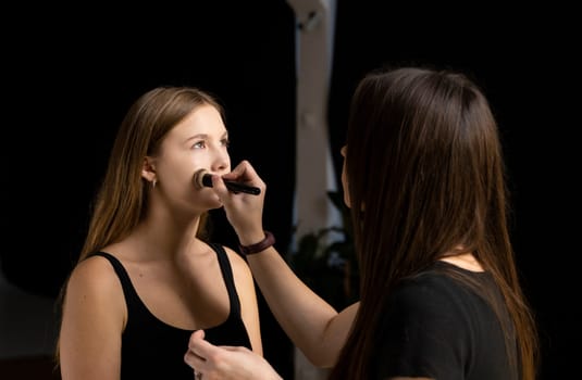 Process of making makeup. Make-up artist working with brush on model face. Portrait of young blonde woman in beauty saloon interior. Applying tone to skin