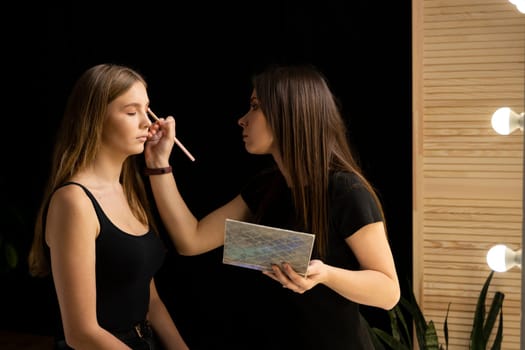 Makeup artist applies eye shadow on a beautiful woman face. Hand of visagiste, painting cosmetics of young beauty model girl. Make up in process