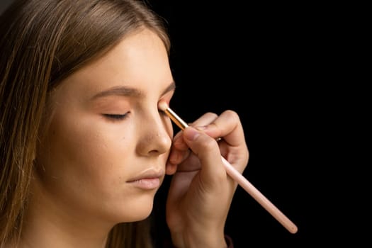 Make-up artist applying eyeshadow in the corner of model eyes and holding a shell with eyeshadow in a black studio. Professional make up