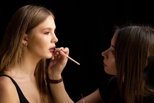 Makeup artist applies red lipstick on a beautiful young woman model face. Hand of make-up master, painting lips of young beauty model girl. Make up in process