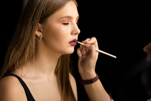 Makeup artist applies red lipstick on a young beautiful woman face. Hand of make-up master, painting lips of young beauty model girl. Make up in process