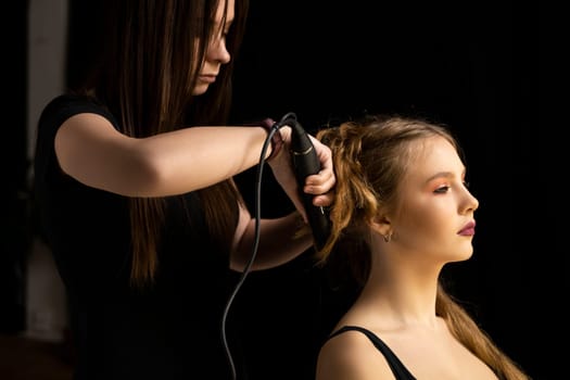 Hair stylist prepares woman makes curls hairstyle with curling iron. Long light brown natural hair